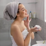 Mulher sorrindo aplicando creme no rosto em ambiente iluminado